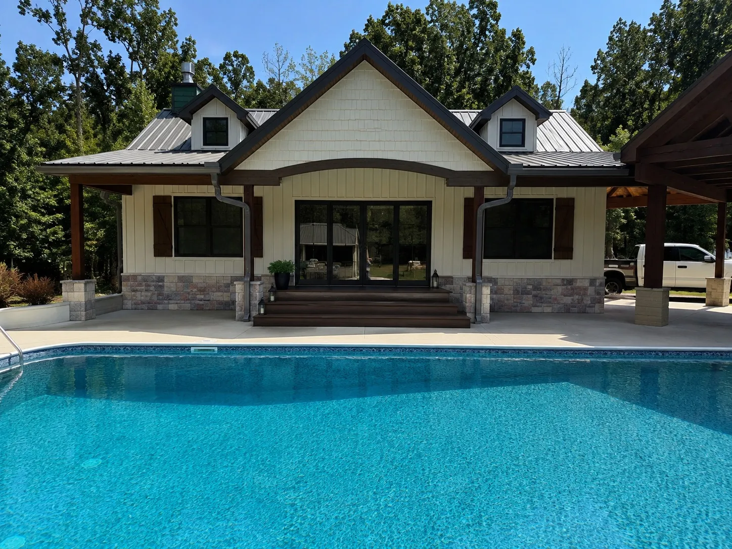 Pool house with board and batten siding, stone wainscot, dormers and wide glass doors overlooking the pool